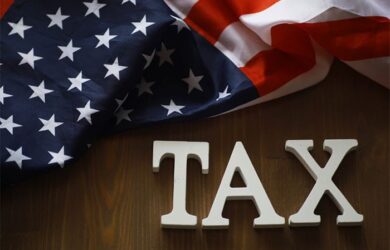 The image shows a wooden surface with white letters spelling TAX and a U.S. flag draped above. The flag displays stars and stripes in red, white, and blue.
