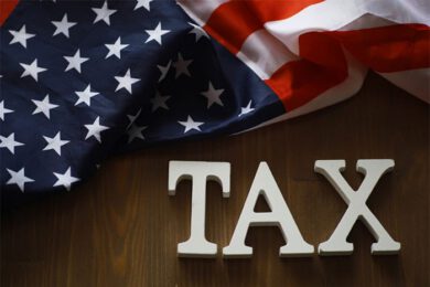 The image shows a wooden surface with white letters spelling TAX and a U.S. flag draped above. The flag displays stars and stripes in red, white, and blue.
