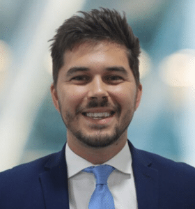 A man in a blue suit and light blue tie smiles at the camera. The background is blurred, with soft lighting.