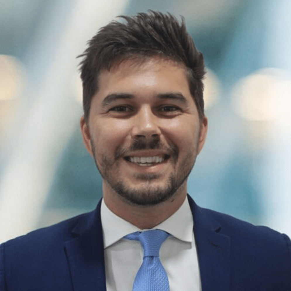 A man in a blue suit and light blue tie smiles at the camera. The background is blurred, with soft lighting.