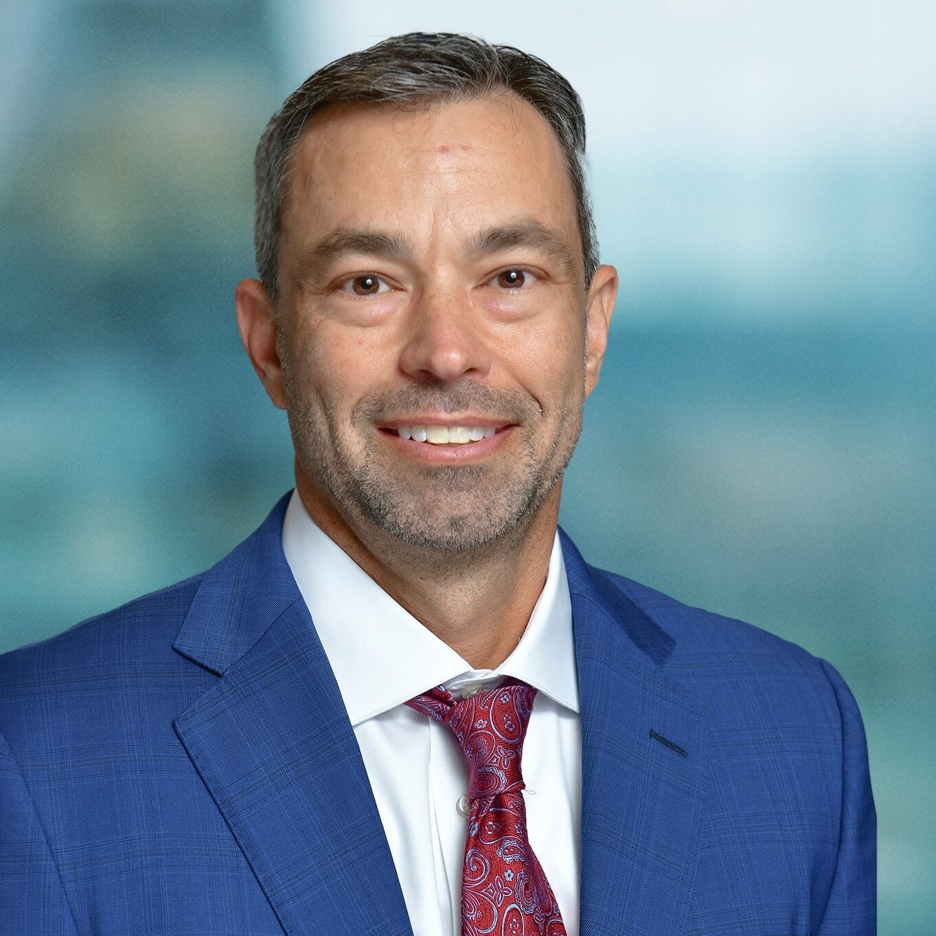 A man in a blue suit and red patterned tie smiles at the camera. He has short brown hair and a light beard. The background is blurred, giving a professional portrait appearance.