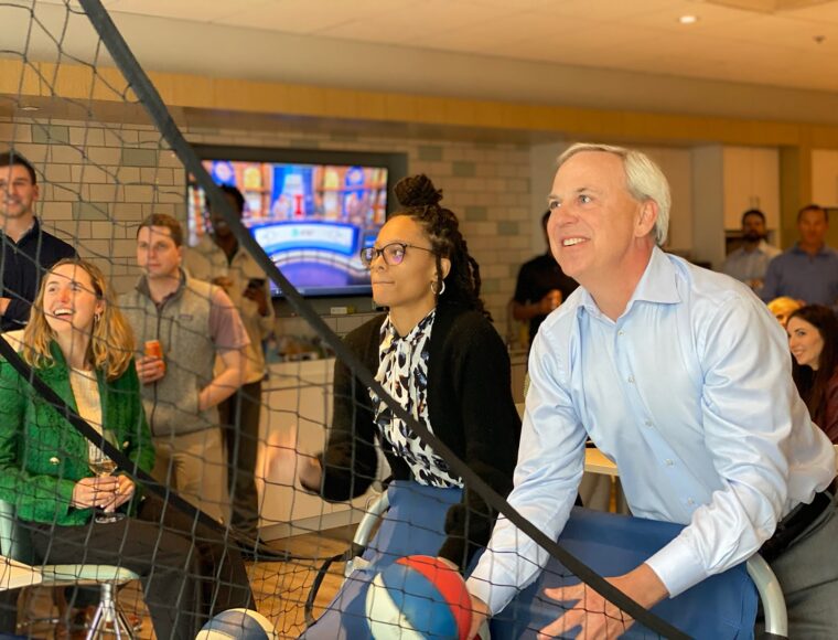 A group of people are enjoying an indoor basketball game. Two individuals are about to shoot basketballs, smiling and focused on the game. Others watch, some holding drinks. A net separates them from the viewer. A TV is visible in the background.