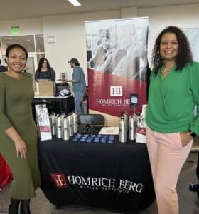 Two women smile in front of a Homrich Berg Wealth Management booth at a career fair. The table displays branded water bottles, pens, and brochures. A large banner with the company logo stands behind them. Other booths and people are visible in the background.