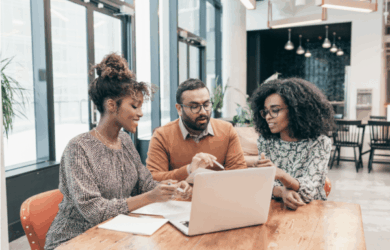 Three people are sitting at a wooden table, engaged in discussion while looking at a laptop. The setting appears to be a modern office with large windows and natural light. There are notebooks and papers on the table.