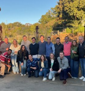 A group of people gathered outdoors, posing for a photo. Theyre casually dressed and standing or kneeling on a paved surface in front of a wooden fence, with trees and a clear blue sky in the background.