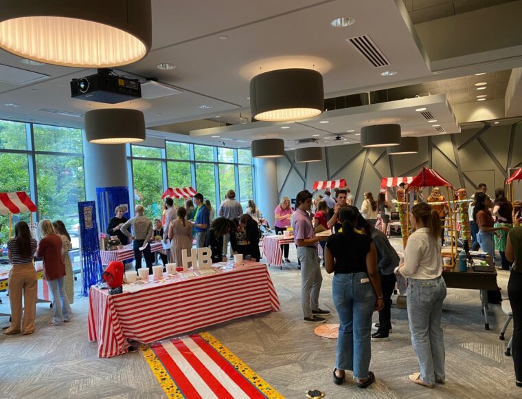People are gathered in a large room with striped red and white stalls, similar to a market or fair. Some individuals are interacting with the stalls while others are mingling. The room is well-lit and has large windows on one side.