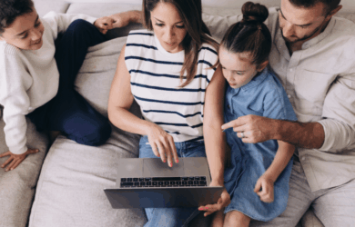 A family of four sits on a couch, gathered around a laptop. The mother points at the screen, while the father and two children, a boy and a girl, look on with interest. They appear engaged and focused on the laptop.