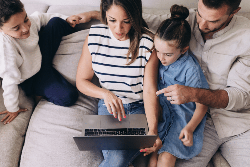 A family of four sits on a couch, gathered around a laptop. The mother points at the screen, while the father and two children, a boy and a girl, look on with interest. They appear engaged and focused on the laptop.