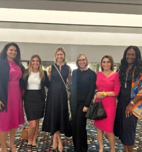 Six women, dressed in a mix of formal and vibrant attire, stand together on a patterned carpet. The group smiles in a bright, modern indoor setting with stripes and geometric lines in the background.