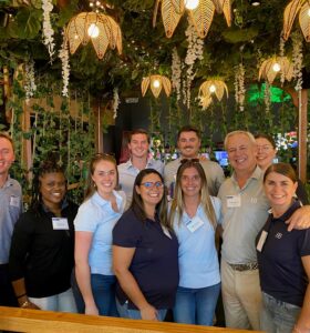 A group of ten people smiling and posing together indoors. The setting features hanging plants and warm lighting. Most individuals are wearing name tags and casual attire. The atmosphere is friendly and relaxed.