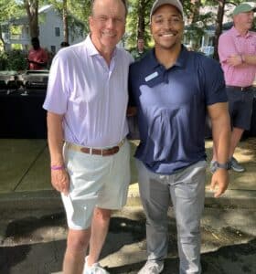 Two men are smiling and posing together outdoors at an event. One is wearing a light purple polo and white shorts, and the other is in a dark blue polo and gray pants. People and trees are visible in the background.