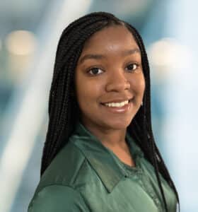 A person with long braided hair smiles, wearing a dark green shirt. The background is softly blurred, featuring a gradient mix of light blue and white tones.