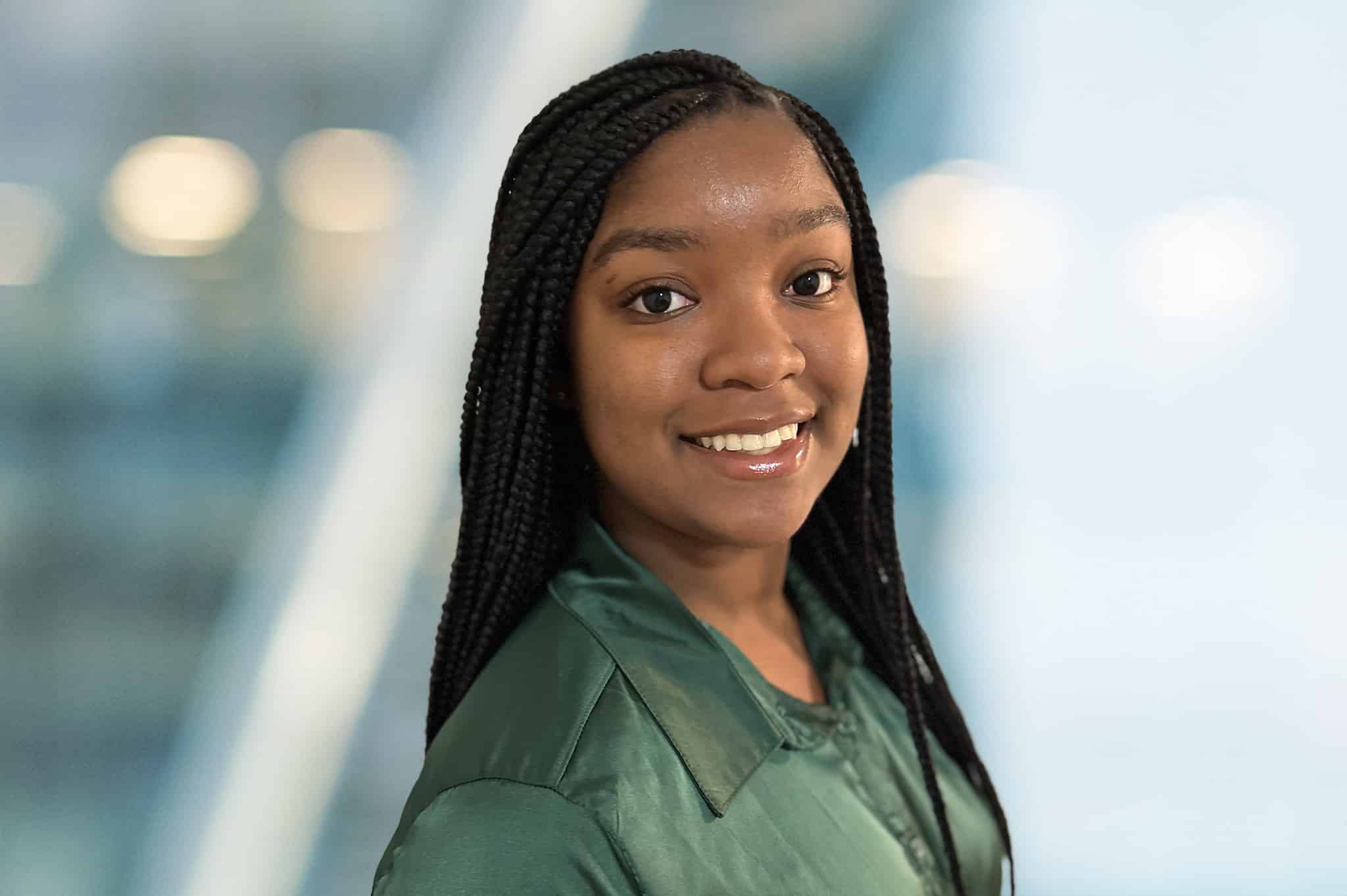 A person with long braided hair smiles, wearing a dark green shirt. The background is softly blurred, featuring a gradient mix of light blue and white tones.