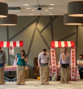 Four people stand in burlap sacks, preparing for a sack race indoors. A carnival-themed setup with striped tents and decor is visible in the background. Onlookers are seated and standing around the room, watching the event.