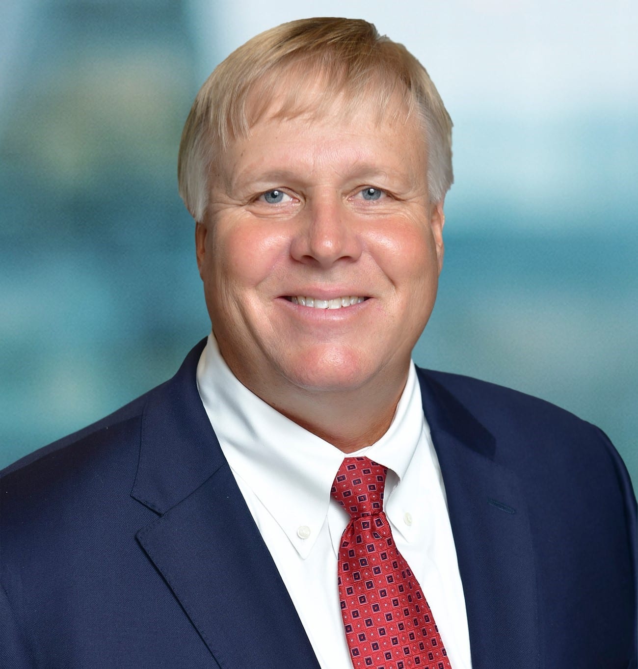 Smiling man in a suit and red tie, with short blonde hair, standing in front of a blurred background.
