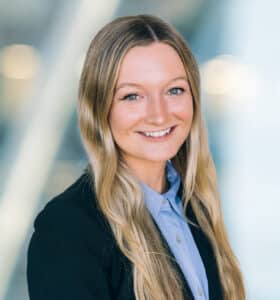 Smiling woman with long blonde hair wearing a black blazer and light blue shirt, standing in front of a blurred background with lights.