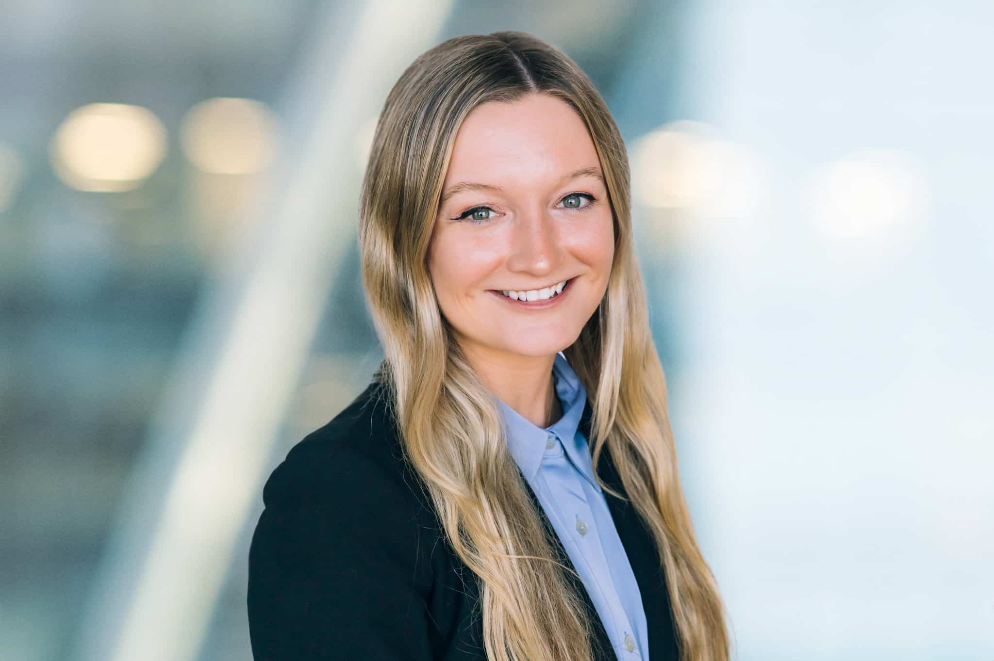Tara Sack Smiling woman with long blonde hair wearing a black blazer and light blue shirt, standing in front of a blurred background with lights.
