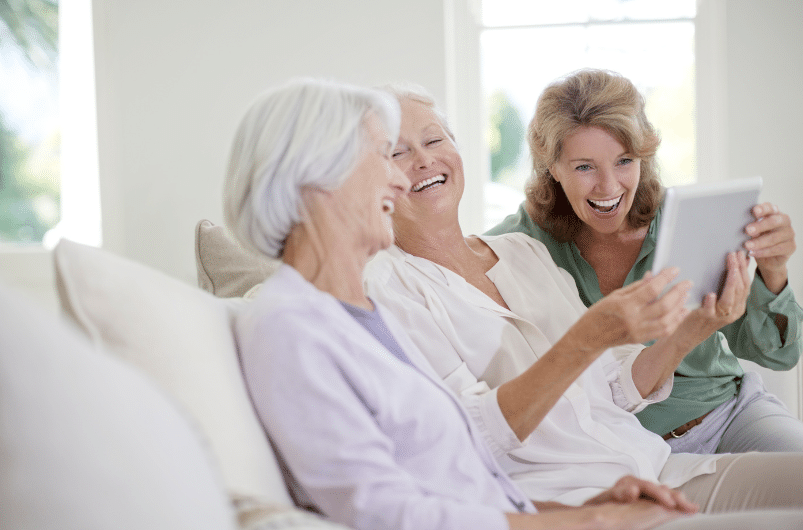 Three women sitting on a couch, enjoying a laugh while looking at a tablet. The woman on the left has short white hair, the middle woman has short blonde hair, and the woman on the right has shoulder-length hair. They are in a well-lit room.
