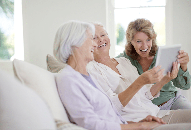 Three women sitting on a couch, enjoying a laugh while looking at a tablet. The woman on the left has short white hair, the middle woman has short blonde hair, and the woman on the right has shoulder-length hair. They are in a well-lit room.