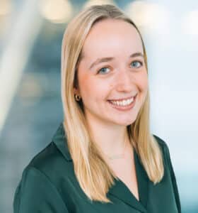 A person with long blonde hair smiles at the camera. They are wearing a dark green shirt and small hoop earrings. The background is softly blurred, featuring light and abstract shapes, suggesting an indoor setting.
