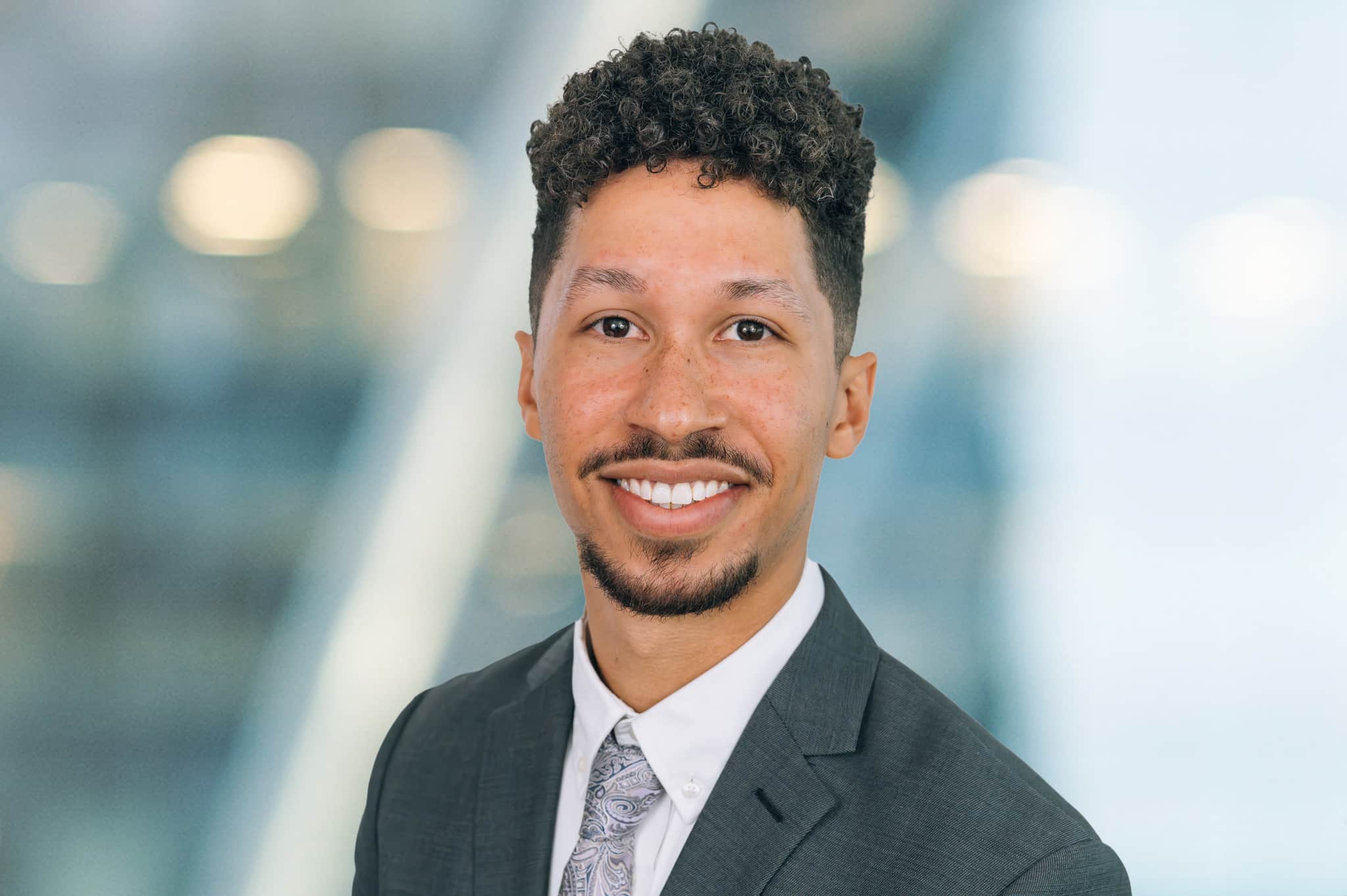 A man with curly hair and a beard smiles while wearing a gray suit and patterned tie. The background is blurred with soft, out-of-focus lights.
