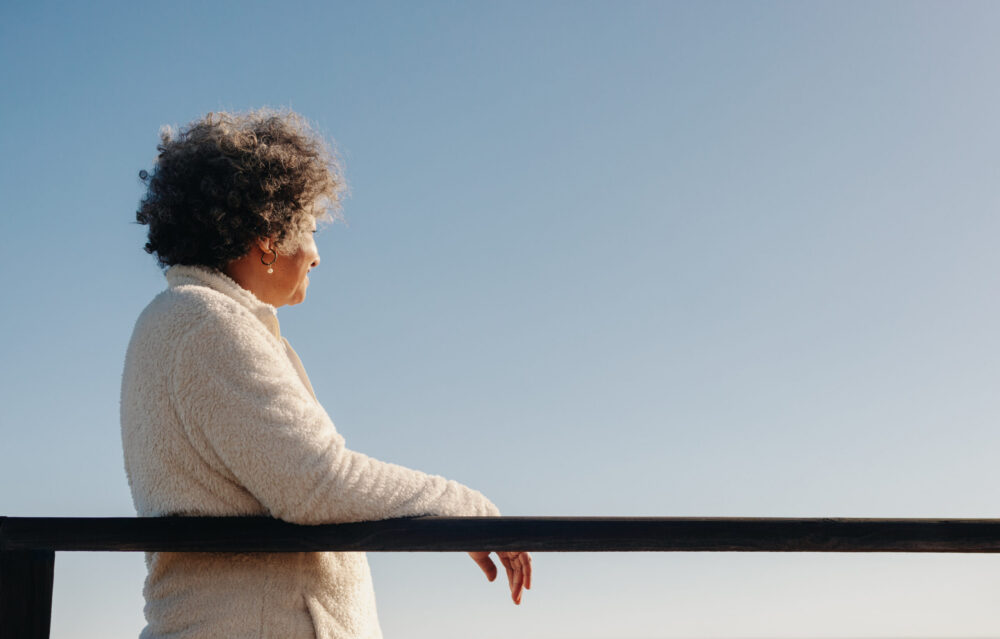 Retired senior woman looking at a view of the ocean while standing on a boardwalk at the beach. Happy elderly woman enjoying a refreshing seaside holiday after retirement.