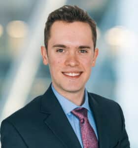A young man wearing a dark suit, blue shirt, and patterned tie is smiling with a blurred background of bright lights.