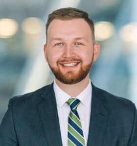 Smiling man with short hair and a beard, wearing a dark suit, white shirt, and green-striped tie, stands against a blurred background with circular light reflections.