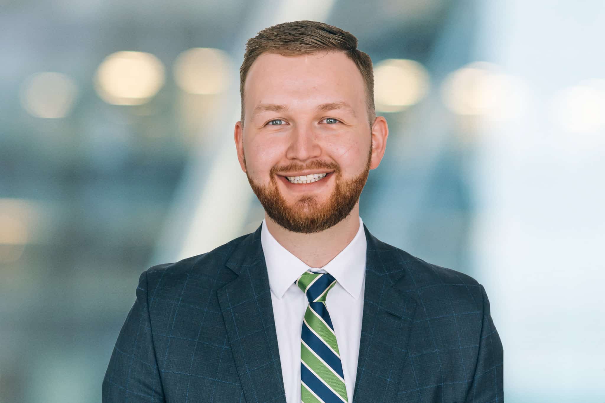 Smiling man with short hair and a beard, wearing a dark suit, white shirt, and green-striped tie, stands against a blurred background with circular light reflections.