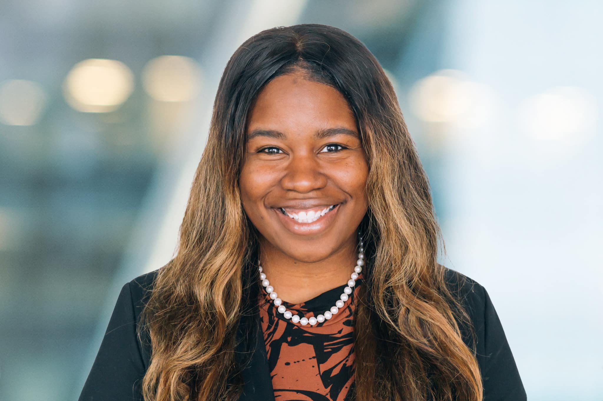 Smiling woman with long, wavy hair wearing a black blazer, a patterned shirt, and a pearl necklace. She is standing in front of a blurred background with lights.