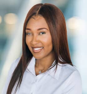 A person with long brown hair smiles, wearing a white shirt and a necklace with a pendant. The background is softly blurred, featuring soft lights and a modern, neutral-toned setting.