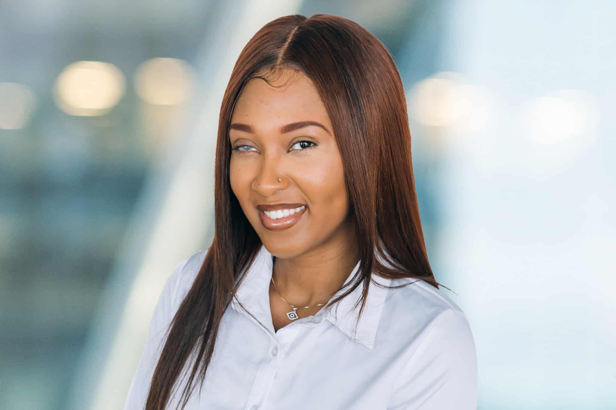 A person with long brown hair smiles, wearing a white shirt and a necklace with a pendant. The background is softly blurred, featuring soft lights and a modern, neutral-toned setting.