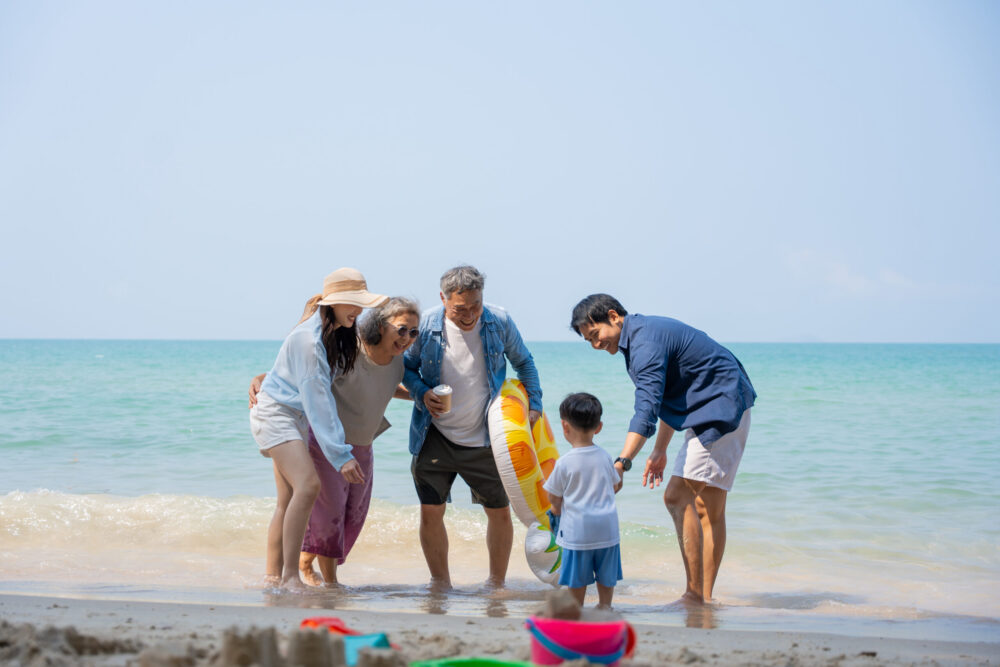 A family of five, including a young child and four adults, stands together at the edge of the sea on a sunny beach. The adults smile and interact with the child, who faces the water, while beach toys lie on the sand.
