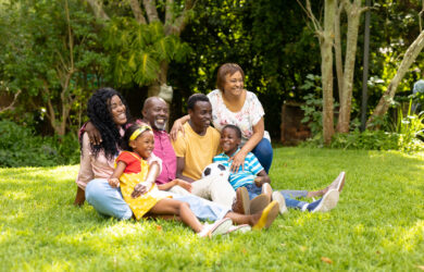 Three generations of a family, including adults and children, sit together on grass in a sunny, green garden, smiling and enjoying each other’s company. Trees and plants are visible in the background.