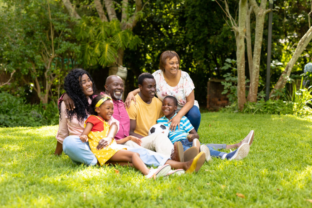 Three generations of a family, including adults and children, sit together on grass in a sunny, green garden, smiling and enjoying each other’s company. Trees and plants are visible in the background.