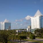 Two modern, glass office buildings with pyramid-shaped tops rise above palm trees and greenery under a blue sky with scattered clouds. The buildings are labeled Daytona Towers.