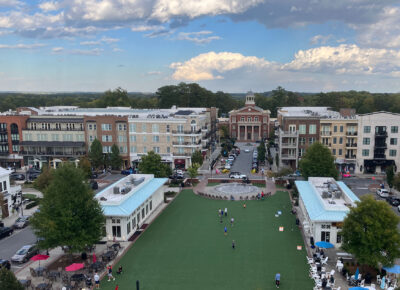 Aerial view of a town square with a large grassy area, people walking and playing, surrounded by restaurants, shops, and multi-story buildings under a partly cloudy sky.