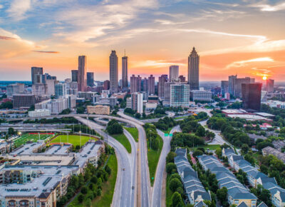 Aerial view of downtown Atlanta at sunset, featuring a colorful sky, tall skyscrapers, highways, and residential areas in the foreground. The sun is partially visible near the horizon.