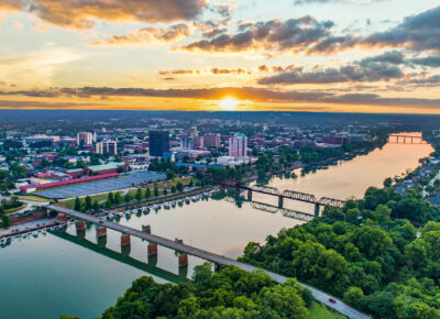 Aerial view of a city at sunset with a river running through it, bridges crossing the water, lush green trees, and buildings spread out under a partly cloudy sky.