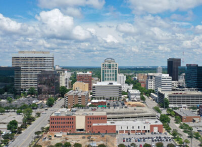 Aerial view of a city downtown with modern high-rise buildings, mid-rise structures, and parked cars under a partly cloudy blue sky. Green trees line the streets between the buildings.