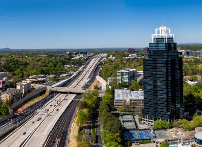 Aerial view of two tall, dark glass skyscrapers beside a busy highway surrounded by trees and mid-rise buildings under a clear blue sky.