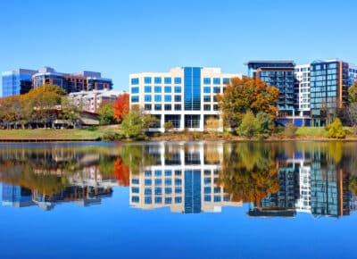 Modern office and apartment buildings with blue glass windows are reflected in a calm lake, surrounded by autumn trees with colorful foliage under a clear blue sky.