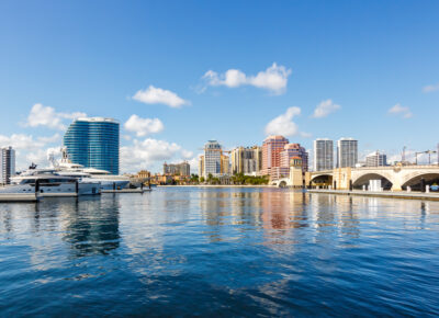 City skyline with modern high-rise buildings, yachts docked along the waterfront, and a stone bridge crossing the water under a bright blue sky with scattered clouds.
