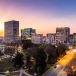 A panoramic view of a city skyline at sunset, featuring historic and modern buildings, including a domed government building, with streets and trees illuminated by streetlights.