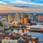 Aerial view of downtown Tampa, Florida at sunset, featuring modern high-rise buildings, waterfront structures, and the Hillsborough River reflecting colorful sky and city lights.