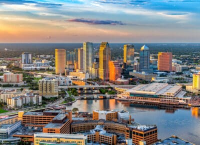 Aerial view of downtown Tampa, Florida at sunset, featuring modern high-rise buildings, waterfront structures, and the Hillsborough River reflecting colorful sky and city lights.
