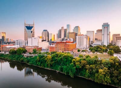 A view of downtown Nashville, Tennessee at sunset, featuring modern skyscrapers, including the AT&T Building, historic brick buildings, and lush green trees along the riverbank with the city skyline reflected in the water.