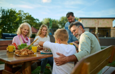 A family of five, including a child, enjoys a meal together outdoors at a wooden table with food and drinks, smiling and interacting in a sunny backyard setting.