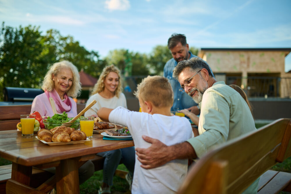 A family of five, including a child, enjoys a meal together outdoors at a wooden table with food and drinks, smiling and interacting in a sunny backyard setting.