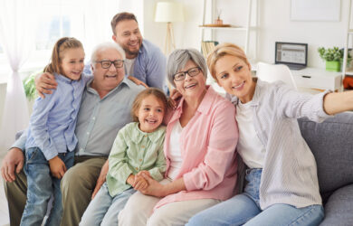 Three generations of a smiling family—two children, two grandparents, and two adults—sit closely together on a sofa, posing happily for a selfie in a bright living room.